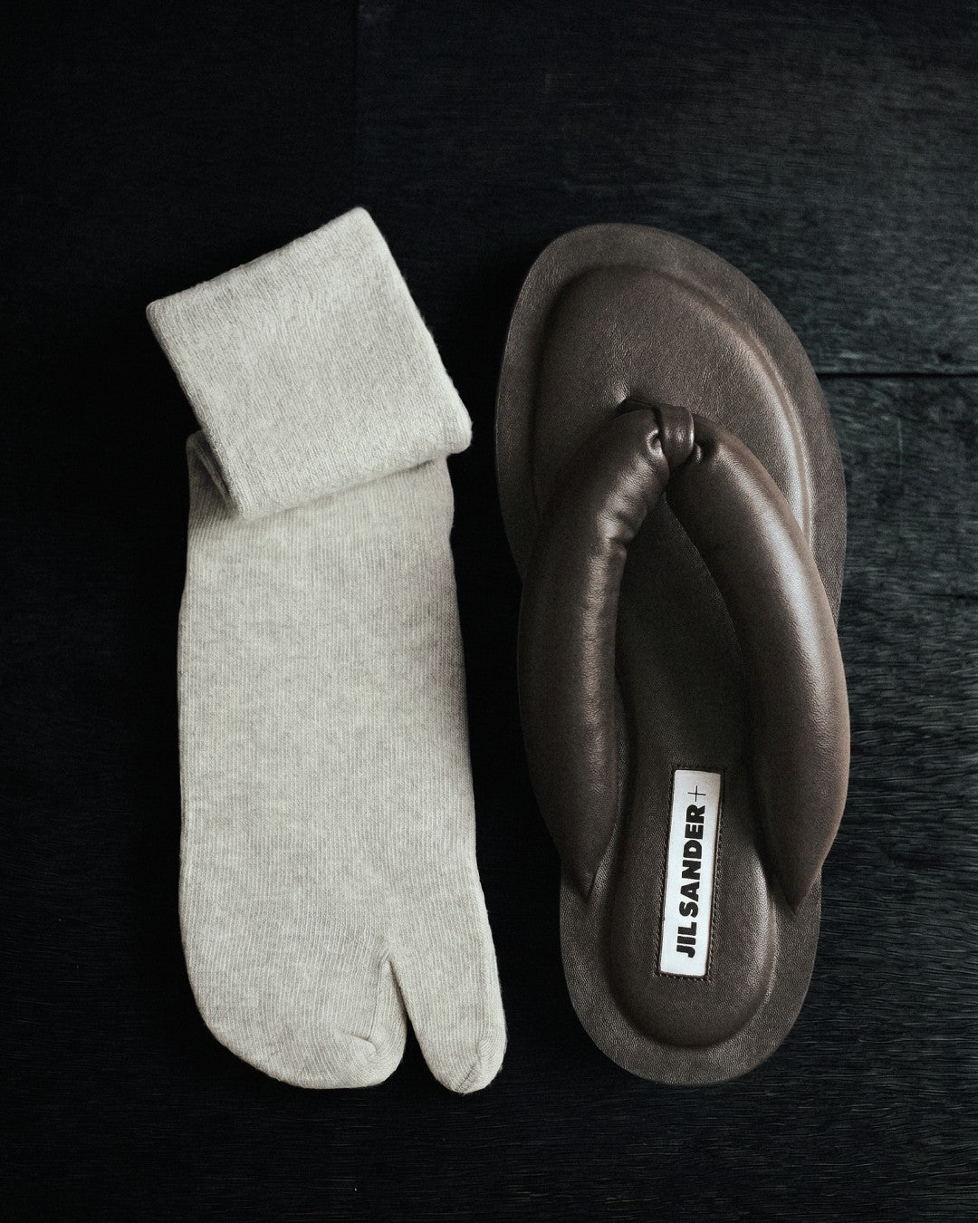SUNMOOL beige fuzzy tabi socks paired with dark brown leather thong sandals on a black wooden surface