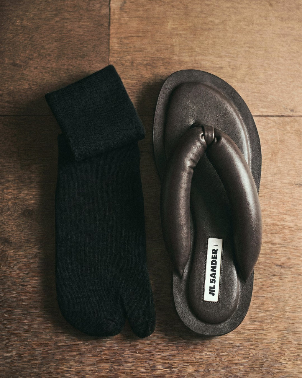 SUNMOOL black fuzzy tabi socks paired with dark brown leather thong sandals on a wooden surface