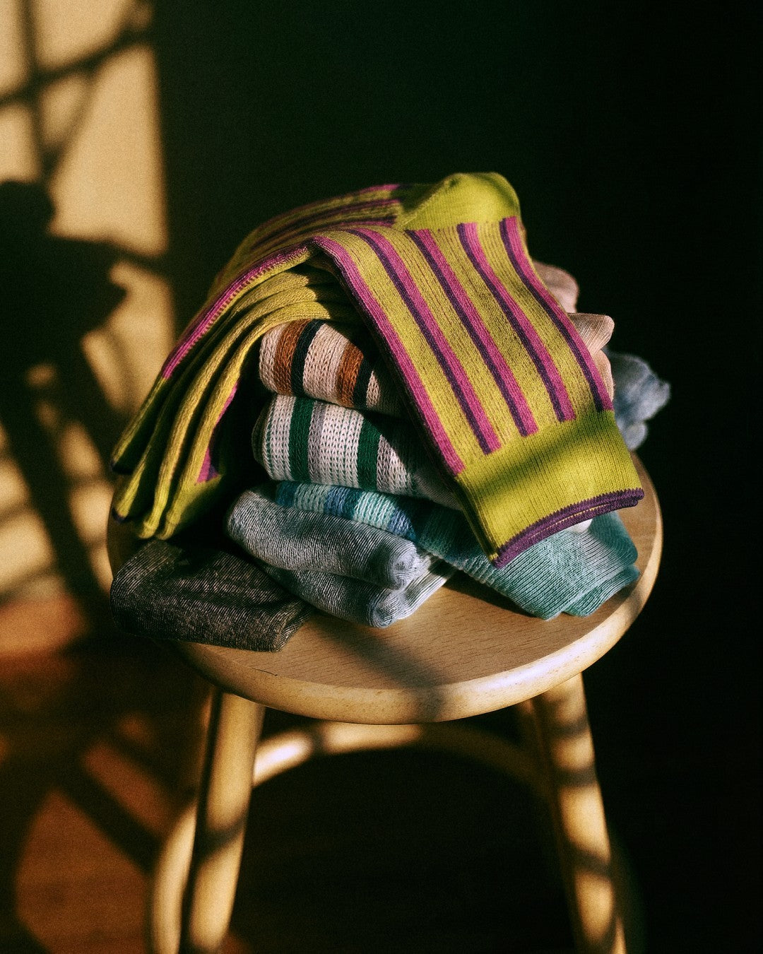 SUNMOOL dual stripe socks in various colors stacked on a wooden stool with warm sunlight and shadows