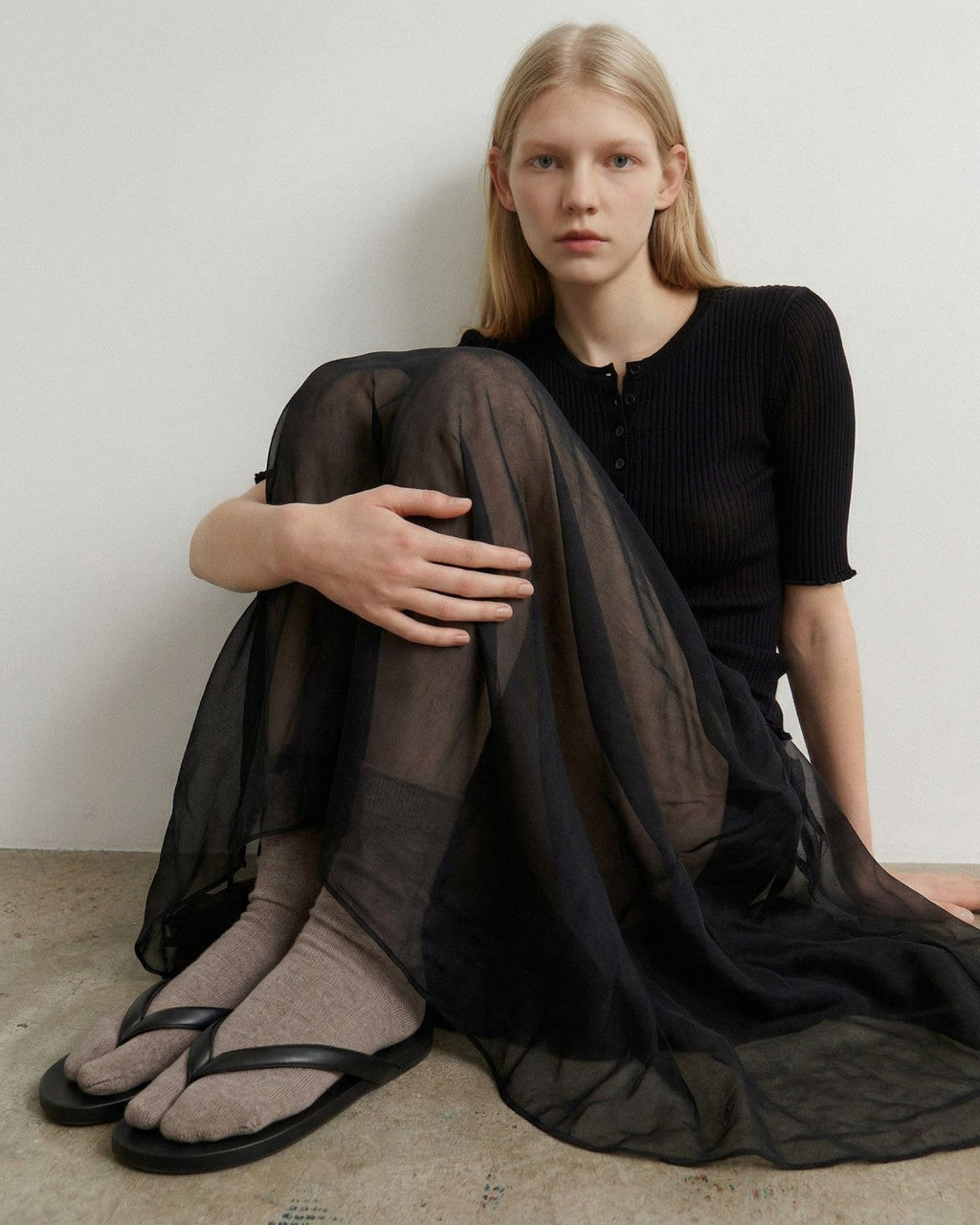 SUNMOOL oatmeal fuzzy tabi socks worn with black thong sandals and a sheer black skirt by a model sitting on the floor